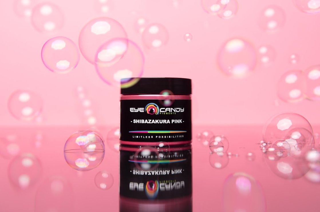 A jar of Eye Candy Pigments Shibazakura Pink sits on a glossy pink surface. Shining bubbles surround the container.